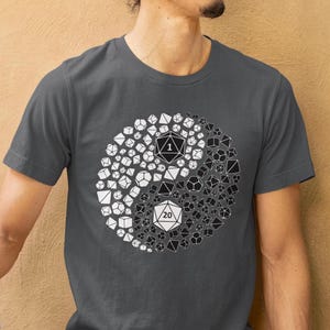 May include: Dark gray t-shirt featuring a yin-yang design composed of dice. The design is split into black and white sections, with dice numbered 1 and 20. The shirt is worn by a person.