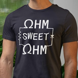 Puede incluir: Camiseta gris oscuro con el texto blanco "OHM SWEET OHM", diseñada para parecerse a un diagrama de circuito eléctrico. La camiseta es de manga corta y está hecha de un material suave.