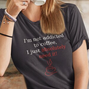 Peut inclure: T-shirt gris fonc&eacute; avec un col en V et le texte "I'm not addicted to coffee, I just absolutely need it!" en blanc et rouge. Le t-shirt pr&eacute;sente un dessin de tasse &agrave; caf&eacute; rouge. La personne porte des lunettes et boit dans une tasse blanche.