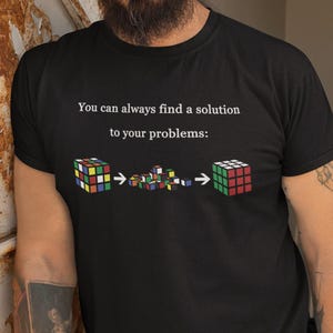 Può includere: Una maglietta nera con una grafica bianca di un cubo di Rubik. La grafica dice "You can always find a solution to your problems:" e mostra un cubo di Rubik in tre fasi di risoluzione.