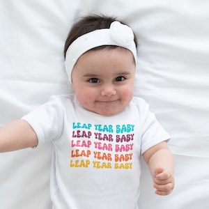 Puede incluir: Un body blanco para bebé con el texto "Leap Year Baby" repetido cinco veces en colores arcoíris.
