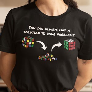 Può includere: Una maglietta nera con una grafica bianca di un cubo di Rubik. La grafica mostra due cubi di Rubik con frecce che puntano verso un mucchio di cubi più piccoli. Il testo "YOU CAN ALWAYS FIND A SOLUTION TO YOUR PROBLEMS" è stampato sopra la grafica.