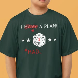 Puede incluir: Camiseta verde oscuro con el texto "I HAVE A PLAN!" tachado en rojo, y "*HAD..." debajo. Un gráfico de dado D20 blanco está centrado, flanqueado por estrellas blancas. La camiseta la lleva una persona.