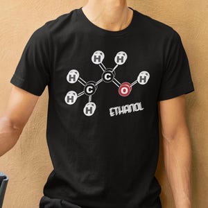 Może przedstawiać: Czarny t-shirt z białym i czerwonym wzorem cząsteczki etanolu. Słowo "ETHANOL" jest wydrukowane poniżej na biało. Osoba nosząca koszulkę ma ciemne włosy.