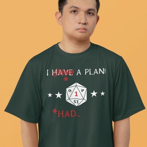 Op de afbeelding: Een donkergroen T-shirt met de tekst "I HAVE A PLAN!" doorgestreept met rode lijnen en de tekst "*HAD..." eronder. Een witte 20-zijdige dobbelsteen met een rode "1" bevindt zich in het midden van het shirt.