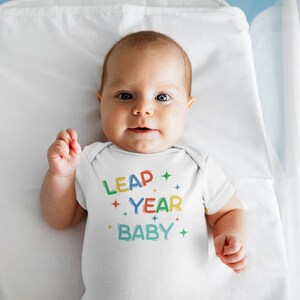 Puede incluir: Un bebé con un body blanco con el texto "Leap Year Baby" en letras de colores y estrellas.