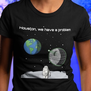 Könnte beinhalten: Ein schwarzes T-Shirt mit einer weißen Grafik eines Astronauten, der auf dem Mond steht und eine Todesstern und die Erde betrachtet. Der Text "Houston, wir haben ein Problem" ist über der Grafik gedruckt.