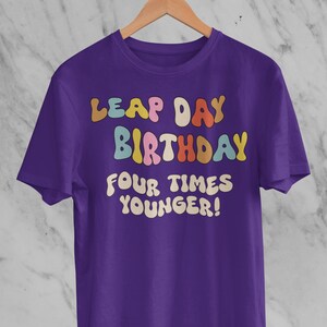 Puede incluir: Camiseta morada con el texto "Leap Day Birthday Four Times Younger!" en letras coloridas y redondas.