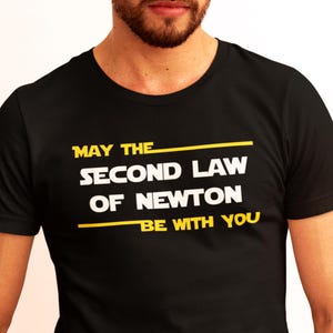 Pode incluir: T-shirt preto com o texto "MAY THE SECOND LAW OF NEWTON BE WITH YOU" em branco e amarelo. A camisa tem gola redonda e mangas curtas.