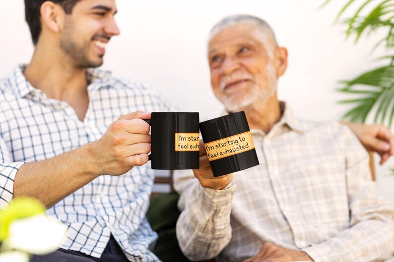 A black ceramic mug held by two people clinking their cups together, featuring a pixel-art style message reading “I'm starting to feel exhausted.” with a low energy bar next to the text. The mug has a cozy farming game aesthetic with an orange-beige text box and pixelated font. Perfect for fans of farming simulation games, cozy gaming and pixel art inspired coffee mugs.
