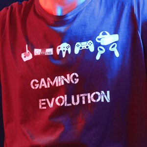 May include: Dark red t-shirt with a white graphic of gaming controllers and VR headset, with the text "GAMING EVOLUTION". The design shows the evolution of gaming controllers from the past to the present.