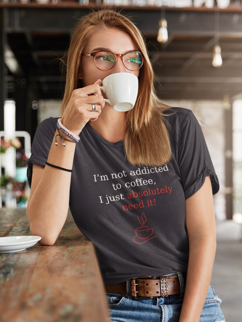 Peut inclure: T-shirt gris fonc&eacute; avec un col en V et le texte "I'm not addicted to coffee, I just absolutely need it!" en blanc et rouge. Le t-shirt pr&eacute;sente un dessin de tasse &agrave; caf&eacute; rouge. La personne porte des lunettes et boit dans une tasse blanche.