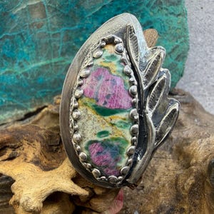 May include: A silver ring featuring a large, teardrop-shaped stone with mottled patterns of magenta, green, and cream. The stone is set in a textured silver bezel with a decorative, leaf-like silver accent extending along one side of the band.