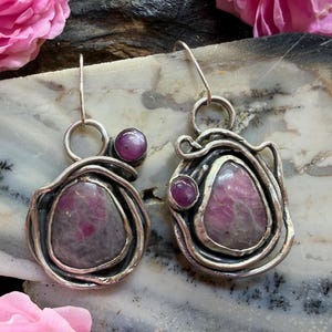 May include: A pair of dangle earrings featuring irregular, polished pink and grey ruby stones set in oxidized silver wire frames. Each earring includes a larger central stone and a smaller, round, deep pink accent stone suspended from a silver hook.