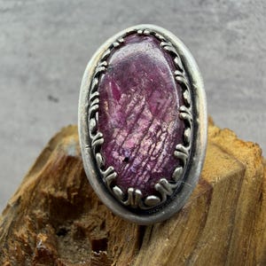 May include: An oval-shaped ring featuring a large, deep magenta gemstone with visible internal crystalline textures. The stone is set in a wide, oxidized silver band with a decorative, scalloped metal border surrounding the central setting.