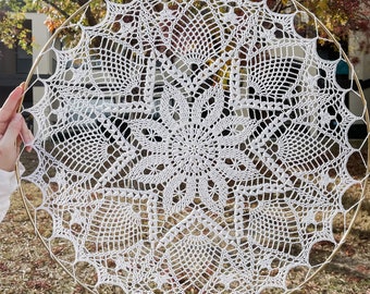 Puede incluir: Un doily de ganchillo blanco con un delicado patrón floral se muestra en un aro de metal dorado.