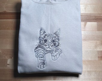 May include: A folded, light gray sweatshirt with a detailed embroidered image of a tabby kitten. The kitten is depicted in shades of gray, with its paws extended. The sweatshirt is made of a soft material.