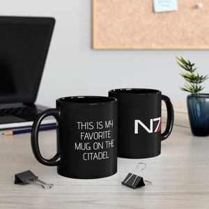 May include: Two black ceramic mugs with white text and a red logo. The mug on the left says "This is my favorite mug on the Citadel". The mug on the right has the red N7 logo.
