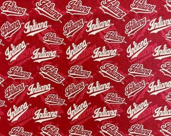May include: A red fabric featuring a repeating pattern of the word Indiana written in a white script font with a trademark symbol. The design is oriented in multiple directions across the surface, creating a dense, uniform graphic print.