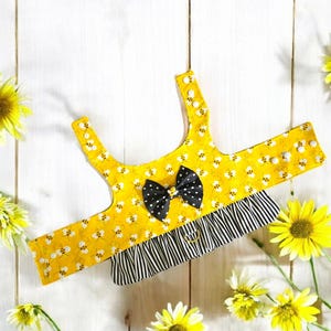 Puede incluir: Arnés amarillo para perro con un lazo negro y estampado de abejas. El arnés tiene un volante de rayas blancas y negras y un anillo plateado.