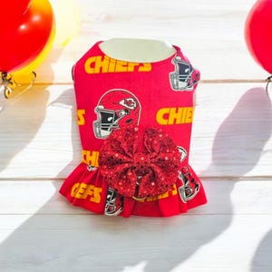 May include: Red dog dress with a red sequined bow, featuring a pattern of football helmets and the word "Chiefs".