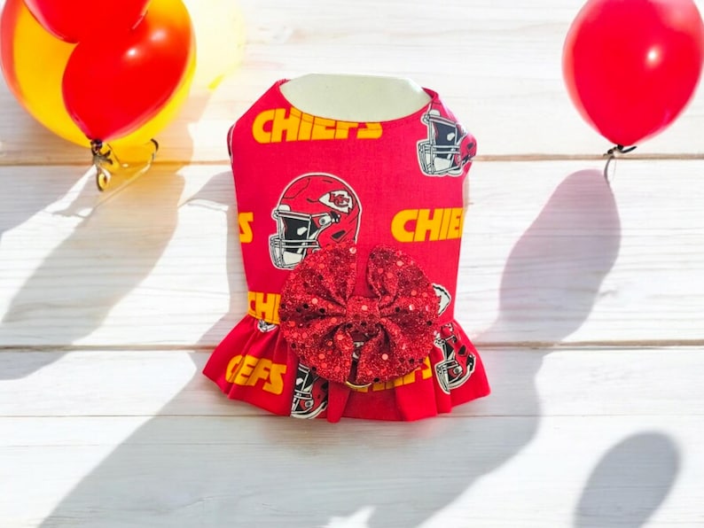 May include: Red dog dress with a red sequined bow, featuring a pattern of football helmets and the word "Chiefs".