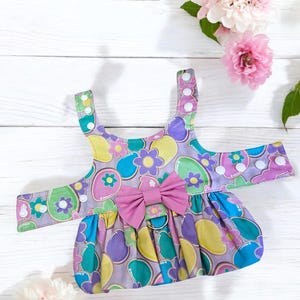 May include: A purple, blue, green, and yellow dog harness with a pink bow. The harness has a floral pattern with a repeating design of colorful eggs.