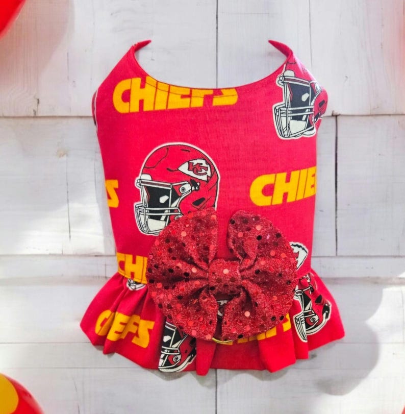 May include: Red and yellow dog dress with a sequined bow. The dress has a pattern of football helmets and the word "Chiefs".