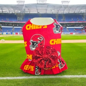 May include: Red and yellow dog dress with a red sequined bow. The dress has a football helmet pattern with the word "Chiefs" repeated on the fabric. The dress is photographed in front of a football stadium.