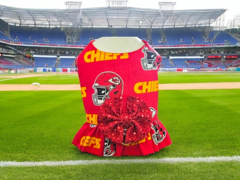 May include: Red and yellow dog dress with a red sequined bow. The dress has a football helmet pattern with the word "Chiefs" repeated on the fabric. The dress is photographed in front of a football stadium.