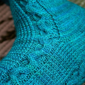 Puede incluir: Primer plano de un calcetín de punto azul turquesa con un patrón de trenza. El calcetín está hecho con un hilo degradado que pasa de un turquesa más oscuro a un turquesa más claro.