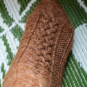 May include: A close-up of a brown knitted sock with a cable knit pattern. The sock is on a green and white patterned surface.