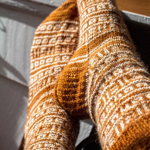 Op de afbeelding: Een paar bruine en witte gebreide sokken met een geometrisch patroon. De sokken zijn gedragen en de voeten rusten op een houten oppervlak. De afbeelding heeft de tekst "Kari Elyssa Designs".
