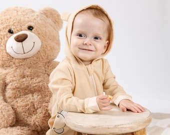 May include: A light beige waffle-knit hooded romper featuring small bear ears on the hood and a front pocket with a bear face graphic. The garment includes a front zipper closure and long sleeves with ribbed cuffs. Text reads: Chanelle is wearing a size 9M.