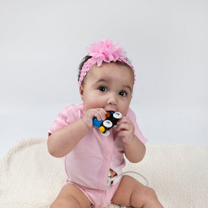 May include: A baby girl wearing a pink onesie and a pink flower headband sits on a white fluffy blanket. She is holding a toy car in her hands and is chewing on it.