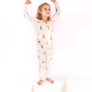 May include: A cream-colored, one-piece pajama set featuring a festive pattern of green and brown Christmas trees and reindeer. The pajamas have a full-length zipper and are worn by a child. Two small decorative Christmas trees are at the child's feet.