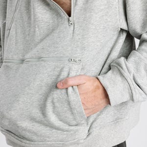 May include: A light gray heathered sweatshirt featuring a diagonal zipper closure at the neckline and a horizontal zippered pocket across the front. The garment has long sleeves with ribbed cuffs and a ribbed hem, with a cream-colored inner lining.