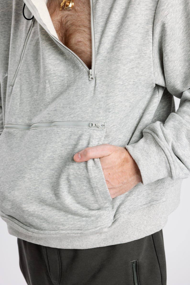 May include: A light gray heathered sweatshirt featuring a diagonal zipper closure at the neckline and a horizontal zippered pocket across the front. The garment has long sleeves with ribbed cuffs and a ribbed hem, with a cream-colored inner lining.