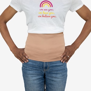 Peut inclure: Une personne portant un t-shirt blanc avec un arc-en-ciel et le texte "we see you. we hear you. we believe you." La personne porte également un gaine de taille beige et un jean bleu.