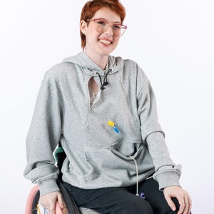 May include: A light gray long-sleeve hooded sweatshirt featuring a front zipper opening and a kangaroo pocket with a zipper closure. The garment includes a medical tube access point. The text Katelyn is 5'6" wearing a size M is visible.