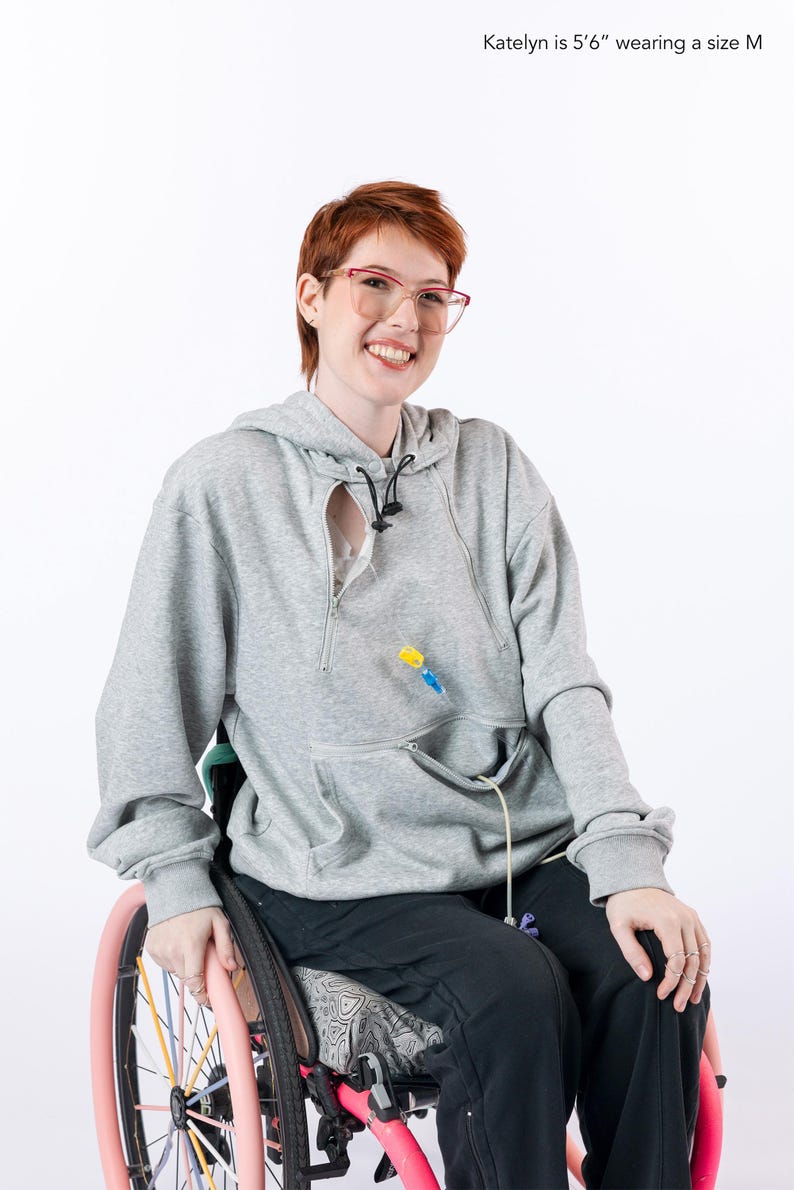 May include: A light gray long-sleeve hooded sweatshirt featuring a front zipper opening and a kangaroo pocket with a zipper closure. The garment includes a medical tube access point. The text Katelyn is 5'6" wearing a size M is visible.