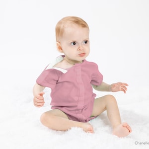 Op de afbeelding: Een baby draagt een roze romper met korte mouwen en een witte rand. De romper heeft een knoopsluiting aan de voorkant. De baby zit op een wit, pluche oppervlak.
