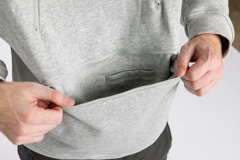 May include: A light gray heathered sweatshirt featuring a large front pocket with a horizontal zipper closure. The garment includes ribbed cuffs and a smaller interior pocket detail with a snap button closure located inside the main zippered pouch.