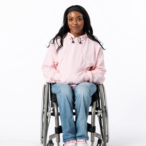 May include: A person in a wheelchair wearing a light pink hoodie, blue jeans, and pink sneakers. The person has long black hair and a black headband. The text at the bottom of the image says "Denisha is 5'5" wearing a size S."