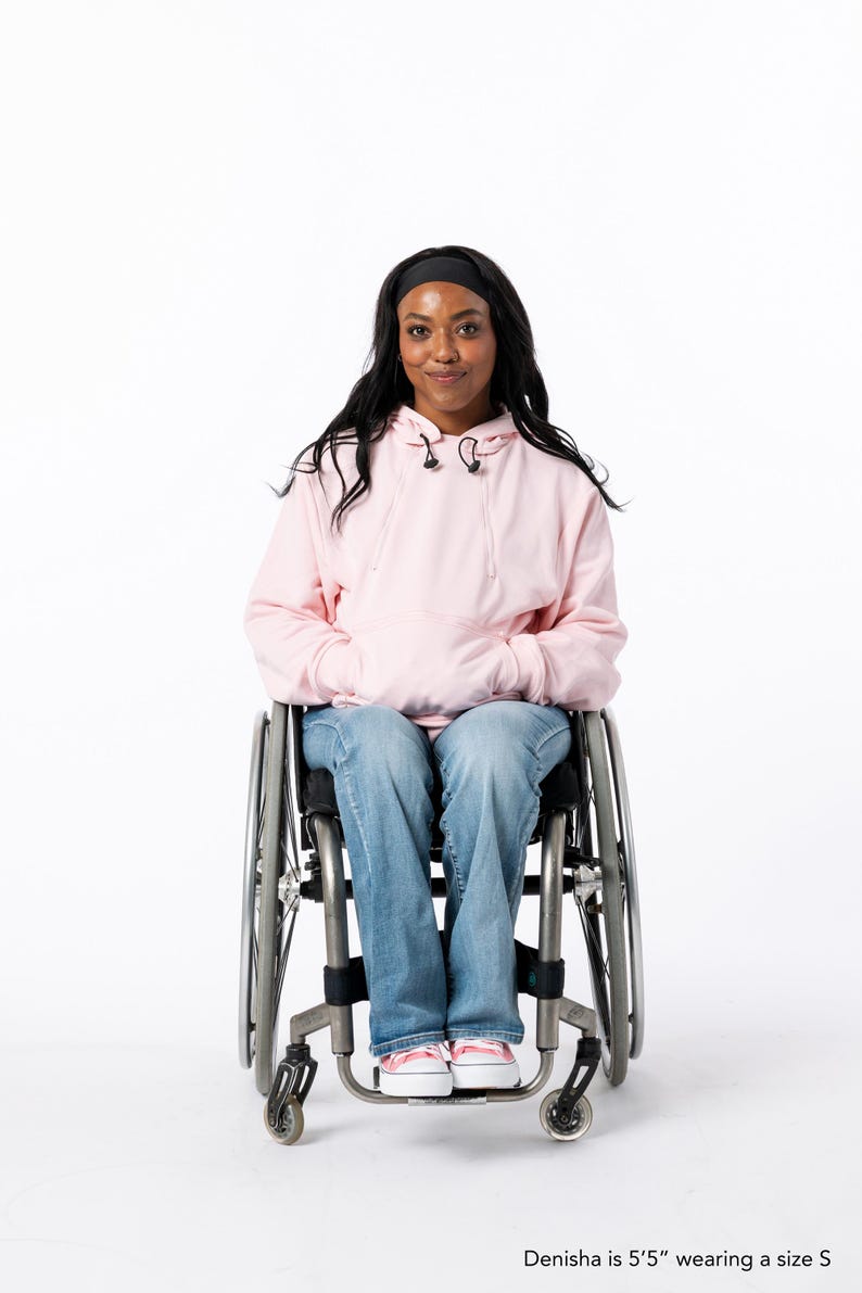 May include: A person in a wheelchair wearing a light pink hoodie, blue jeans, and pink sneakers. The person has long black hair and a black headband. The text at the bottom of the image says "Denisha is 5'5" wearing a size S."
