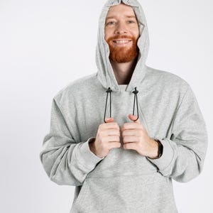 May include: A light gray long-sleeve hooded sweatshirt featuring a large front pocket with a horizontal zipper closure. The hood includes black adjustable drawstrings with plastic toggles, and the garment is constructed from a soft, heathered fabric.