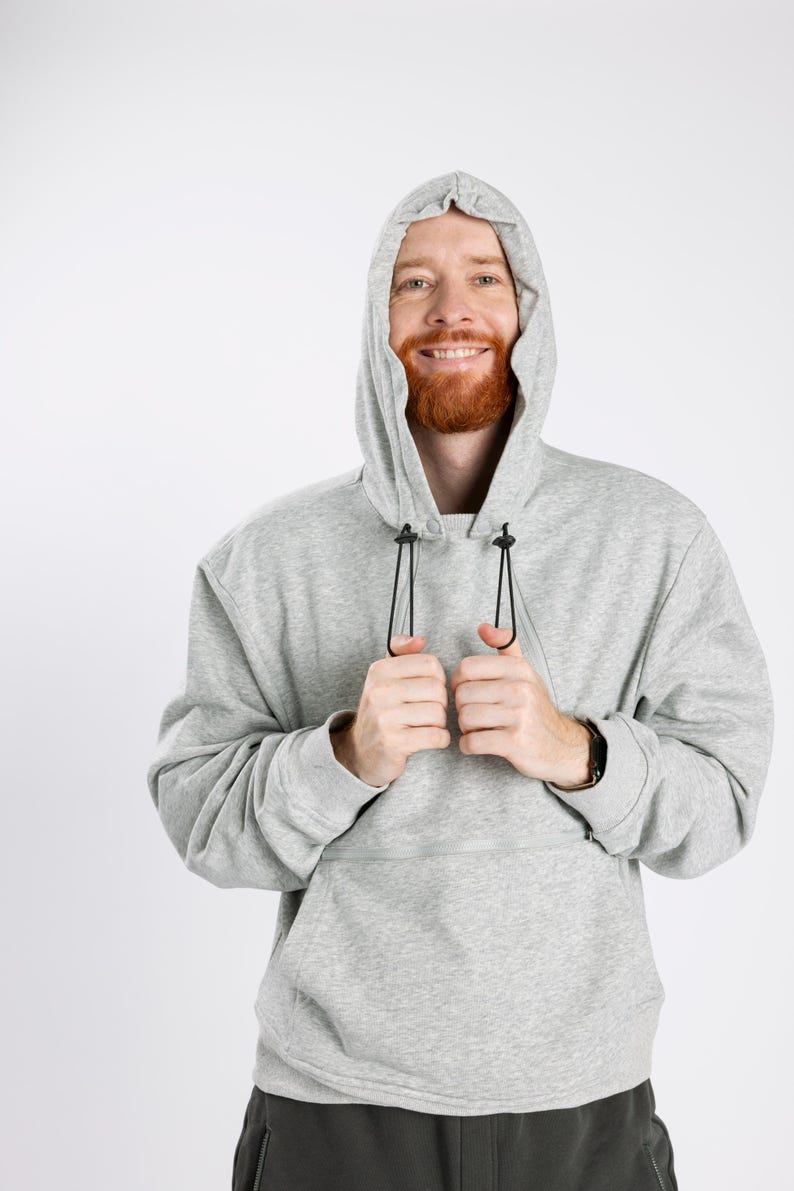 May include: A light gray long-sleeve hooded sweatshirt featuring a large front pocket with a horizontal zipper closure. The hood includes black adjustable drawstrings with plastic toggles, and the garment is constructed from a soft, heathered fabric.