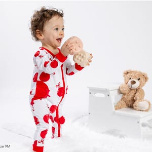 Op de afbeelding: Een witte romper met lange mouwen, een rode appelprint en een rode ritssluiting, gedragen door een peuter die een beige, getextureerd blokspeeltje vasthoudt, met een bruine teddybeer op een wit opstapje. Tekst: Gavin is wearing a size 9M.