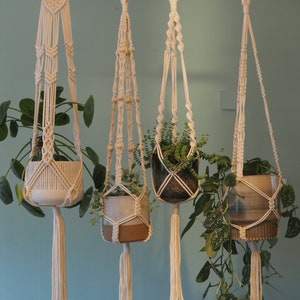 Könnte beinhalten: Vier Makramee-Pflanzenhänger mit Holzperlen und Quasten, die jeweils eine Topfpflanze halten. Die Hänger sind aus natürlichem weißem Baumwollschnur hergestellt und hängen von der Decke.