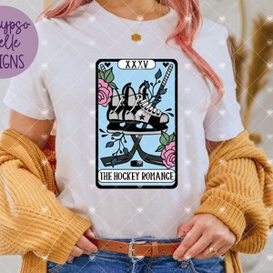 May include: A white t-shirt with a blue and pink tarot card design featuring a pair of hockey skates and sticks with the text "The Hockey Romance" and "XXXV".